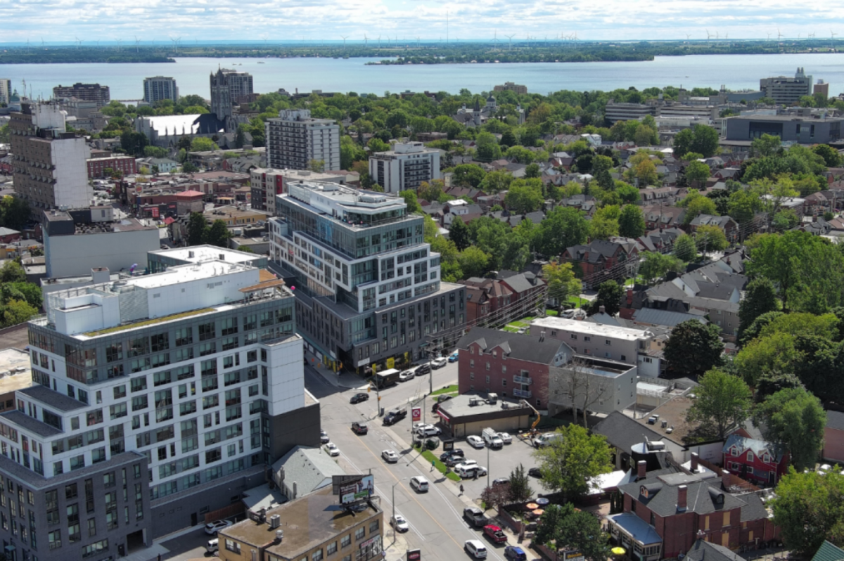 Aerial view of modern mid-rise apartment buildings in downtown Kingston, Ontario, with Lake Ontario and tree-lined neighborhoods in the background.